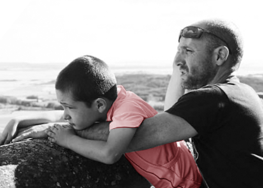 A black and white photo of a boy and a man on a beach leaning on a rock together with the boy’s shirt in red.